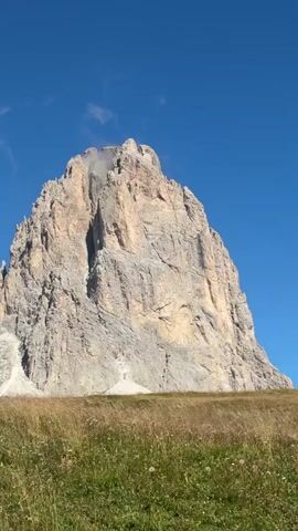 The video shows a panoramic view of a mountain range under a clear blue sky. In the foreground, there is a field of tall, dry grass with some green patches. The mountains are rugged and rocky, with sheer cliffs and sharp peaks. The scale of the mountains is vast, suggesting a remote and wild landscape. There is no movement or activity depicted in the video, other than the slight rustling of the grass and the movement of a few wisps of clouds. The lighting indicates it is daytime, likely late morning or early afternoon, given the brightness and the direction of the shadows.