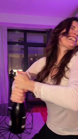A woman in a long-sleeved shirt successfully opens a bottle of sparkling wine with a dramatic flourish in a room with purple lighting. The cork flies off and she holds the bottle up as people in the room cheer. The room appears to be a living room with a window, a coffee table, and people sitting on a sofa. Other people are visible in the background, some recording with their phones. The room also contains wall decorations.