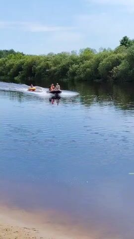 The media file shows a sunny day with a bright blue sky and fluffy white clouds. A wide, calm river flows through a lush green landscape, with dense trees lining the far bank. In the foreground, a sandy riverbank is visible.

The main activity involves a motorboat pulling an inflatable tube on the water. Three people are seated in the motorboat, wearing life vests. Another person is riding on the yellow inflatable tube, also appearing to wear a life vest. The motorboat is creating a white wake as it moves across the water, pulling the tube along. The scene suggests a recreational activity, likely a fun day out on the river.

The overall atmosphere is one of leisure and enjoyment. The bright sunlight indicates it's daytime, probably late morning or early afternoon. The clear sky and green foliage suggest pleasant weather, ideal for water sports. The location appears to be a natural, somewhat remote area, given the extensive tree cover and the absence of buildings or other man-made structures in the immediate vicinity. The water is calm and reflects the sky and surrounding greenery. There is no visible text in the media file.