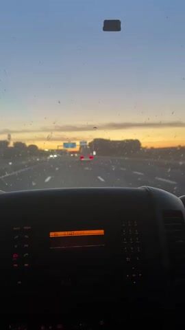 The video shows a view from inside a vehicle, looking out through the windshield at a highway during sunset. The windshield is dirty with visible specks and water spots. The road ahead is visible with multiple lanes and other vehicles, including cars and a large truck. There are overhead signs with blue and white text and arrows indicating directions. The sky is a blend of orange, yellow, and blue, characteristic of a sunset. The dashboard of the vehicle is partially visible at the bottom, showing a radio display and buttons. The overall impression is of a journey at dusk on a highway.