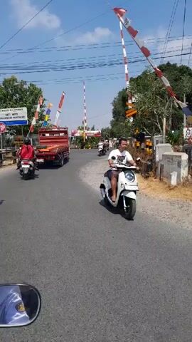 The video begins with a close-up of a motorcycle's side mirror, reflecting a blue sky with white clouds. The perspective then shifts to a first-person view from a motorcycle traveling on a paved road. On the left side of the road, a red dump truck is parked, with a person wearing a red jacket and helmet on a white motorcycle in front of it. Further ahead, several other motorcycles are visible, some with passengers. Red and white striped barriers, typical of a railway crossing, are present on both sides of the road. A sign with Indonesian text is visible on the left, and a building with a sign that reads "SWANT KETABANG" is on the right. The road has some gravel and patches of rough terrain on the right shoulder. The video pans slightly to the right, showing more of the railway crossing and surrounding greenery, including large trees. The sun is shining brightly, casting shadows. The overall scene depicts a moderately busy road at a railway crossing on a clear day.
