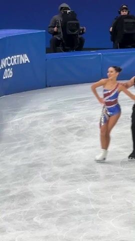 A short video clip shows a pair of ice dancers on a rink. The woman wears a dress patterned with the Union Jack, while the man is in a leopard-print top and dark trousers. They execute a spin, hand-in-hand, before separating. He performs a brief solo spin and skate, and then they rejoin, with the man supporting the woman as she performs a brief, synchronized step. A cameraman and the "MILANOCORTINA 2026" signage is visible in the background.