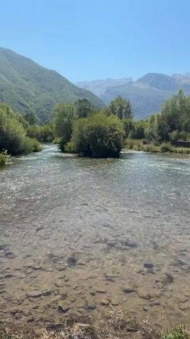The video pans across a clear river with a rocky bottom under a bright blue sky. In the background, lush green trees and rolling mountains are visible. A small, rustic wooden bridge crosses the river in one section of the video, with a few people walking across it. The riverbanks are lined with grass and some scattered foliage. The overall scene depicts a serene natural landscape with elements of human presence.