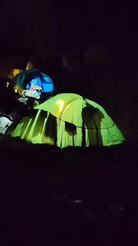 A green tent is illuminated by a light source inside, casting shadows. Behind it, a blue tent is visible in the dark. The tents appear to be set up in an outdoor environment with rocks and dark ground. The scene is dark, suggesting it is nighttime or a poorly lit area.