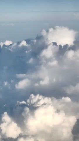 The video shows a high-angle aerial view of a sky filled with clouds. The clouds are white and gray, with some areas brightly lit by the sun. The sky is a pale blue, and the clouds appear to be moving slowly. The overall impression is one of vastness and natural beauty.