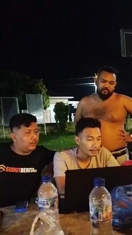 Three men are gathered outdoors at night. One man, wearing a black t-shirt with white text that appears to read "SUDUTBERITA", is seated on the left. Another man is seated in the center, looking at a laptop. He is wearing a grey t-shirt. A third man, shirtless and with dark, curly hair on his chest, stands behind the seated men, with his right hand on his hip. In the foreground, on a wooden table, are two plastic water bottles, one labeled "AQUA", and some other items that are not clearly discernible. The background is dark, suggesting it is nighttime, with some lights and vegetation visible.