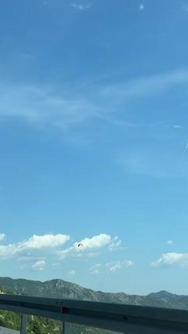 A paraglider is visible in the sky above a mountainous landscape. A speed limit sign for 60 is also visible on the right side of the frame. The sky is blue with scattered clouds. The video appears to be shot from a moving vehicle.