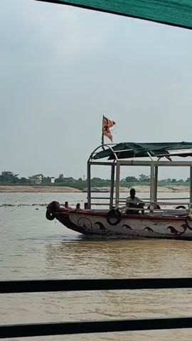 A boat with a canvas covering sails on a murky river. A person sits at the helm of the boat, and another person stands at the bow. The boat has a decorative design along its side and tires attached to its exterior. A flag is visible on the back of the boat. The riverbanks are lined with vegetation and buildings in the distance. The sky is overcast.