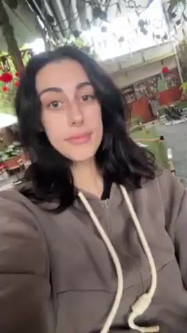 A woman with dark hair is in the foreground, wearing a brown zip-up hoodie. She smiles and makes a kissing face at the camera. Behind her is an outdoor seating area with green chairs and tables. There are hanging red decorations and potted plants. The setting seems to be a cafe or restaurant patio.