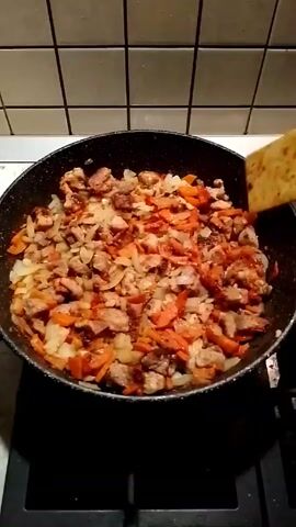 A high-angle, static shot shows a person stirring a frying pan with a wooden spoon. The pan is on a gas stovetop with a light-colored tile backsplash. The pan contains pieces of meat, onions, and what appears to be carrots, all cooking together. The person’s hand is visible while stirring, and the food appears to be well-combined and cooking evenly.