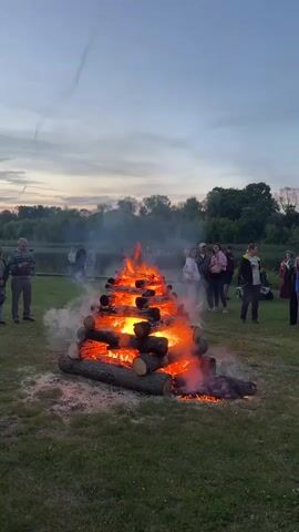 The media file shows an outdoor scene with a large bonfire as the central object. The bonfire is constructed from several layers of logs stacked in a pyramid shape, and it is actively burning with bright orange flames and emitting smoke. Around the bonfire, a group of people are gathered, observing the fire. They appear to be casually dressed, suggesting a relaxed event.

The setting is a grassy field, with a body of water, likely a lake or river, visible in the background. Trees and vegetation line the far bank of the water. The sky is overcast with hints of sunset colors, indicating it is late evening or dusk. The overall mood seems to be one of communal gathering and enjoyment.

The people in the video vary in age, and their presence suggests a social event or celebration. Some are standing, while others are sitting or observing from a distance. There is no visible text in the media file. The time of day, the bonfire, and the gathering of people point towards an evening event, possibly a festival, outdoor party, or a specific cultural celebration. The presence of trees and water suggests a natural, possibly recreational area.