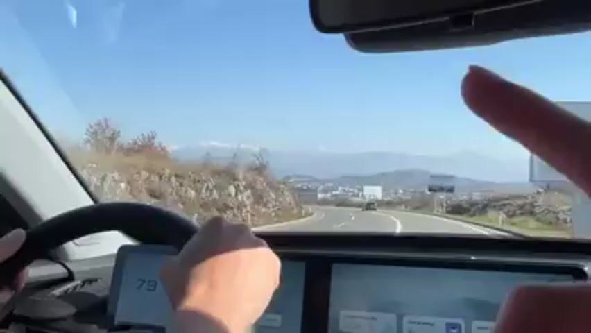 From the driver's seat perspective, the video shows a car driving on a winding road towards a city. Mountains are visible in the distance. There is a billboard on the right side of the road. The car's dashboard shows a speed of 78. Other cars can be seen on the road ahead. The sky is clear and blue. The driver's hands are on the steering wheel.
