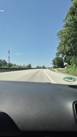 The video is taken from the perspective of a passenger inside a car traveling on a highway. The dashboard is visible in the foreground. The road is paved and has lane markings. On the right side of the road, there is a green circular sign with an arrow indicating a turn. Trees and vegetation line the sides of the highway. The sky is clear and blue, suggesting a sunny day. There are other cars visible in the distance on the highway. The audio contains speech in Russian, where the speaker mentions being on the right and waiting for someone.