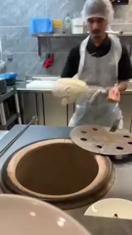 The video shows a person preparing flatbread inside a tandoor oven in a restaurant kitchen. The person, wearing a hairnet and a plastic apron, is placing the flatbread dough into the hot tandoor. The tandoor is a large, round oven with a metal lid with holes. The kitchen has stainless steel counters, gray tile walls, and other equipment. The person is focused on the task of putting the dough into the tandoor oven.