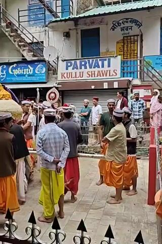 A vibrant procession moves down a narrow street in Kasol, India, featuring a large group of men. Most are dressed in traditional Himachali attire, including distinct white caps with colorful bands and bright dhotis in yellow, orange, and white. Several men are carrying a yellow, decorated palanquin-like structure, likely a deity or sacred object. Many of the carriers are barefoot.

The procession takes place during daytime under clear skies. In the background, multi-story buildings line the street, housing various businesses. Visible signs include "PARVATI KULLU SHAWLS," "G Gobind," and "PADHA FAMILY GUEST HOUSE." Some onlookers are visible on the balconies of the buildings. The scene depicts a cultural or religious event common in the Himachali region.