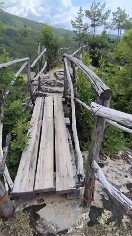 The video shows a POV perspective of someone walking on a narrow wooden bridge with a rustic wooden railing. The bridge is surrounded by lush green foliage and rocky terrain. In the background, there are rolling hills covered in trees and a cloudy sky with patches of blue. The person is walking towards a rocky path that continues beyond the bridge, also bordered by the same wooden railing. The environment appears to be a natural, mountainous landscape.