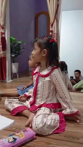 A video shows two young girls sitting on a wooden floor, facing away from the camera. The girl in the foreground is wearing a light-colored pajama set with pink trim and patterns. She has her hair pulled back in two pigtails with red ribbons. The second girl, partially visible behind her, is wearing a blue top and is sitting cross-legged. A pencil case with Disney characters is on the floor in front of the girls. In the background, a mirror reflects a man sitting in a chair and looking at his phone. A potted plant and curtains are also visible in the room. The lighting suggests it is evening.