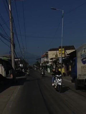The video captures a first-person perspective from a moving vehicle on a sunny day, showcasing a street with moderate traffic, including motorcycles and people. The street has distinct black tire marks, and buildings line both sides. Electrical wires crisscross overhead, attached to wooden and metal poles. The sky is clear blue.