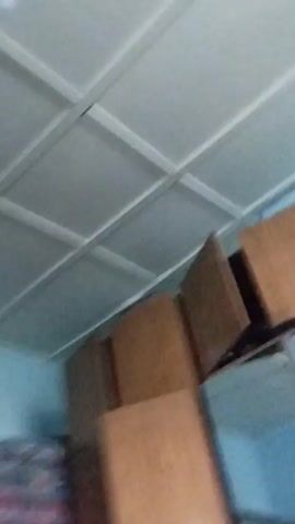 The video shows a room with a ceiling, walls, and furniture. The walls are painted blue. There is a wardrobe with wooden doors and mirrored panels. On top of the wardrobe, there are cardboard boxes. Clothes, including a white shirt, a black jacket, and a cap, are hanging on the wall near the wardrobe. A bed is visible with blankets and pillows. The room appears to be cluttered.