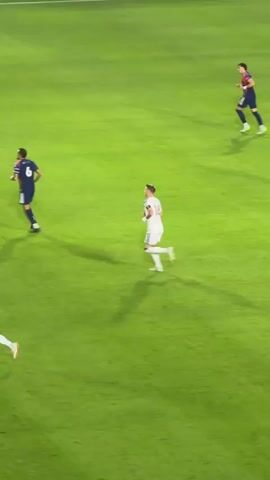 The video shows two soccer players running on a green field. The player on the left is wearing a dark blue jersey with maroon trim and the player on the right is wearing a white jersey with maroon trim. They are both running towards the right side of the frame. The stadium is visible in the background, with spectators in the stands.