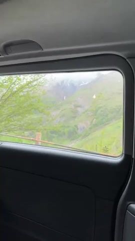 From inside a vehicle, the view through a window reveals a scenic landscape. Initially, the view is obscured by foliage, but as the vehicle progresses, a mountain range comes into view. The mountains are a mix of lush green terrain and rocky areas, with some snow visible at the peaks. The sky is overcast with fog and clouds. A guardrail runs along the side of the road.