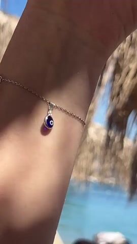 A close-up shot focuses on a wrist with a silver bracelet and a blue evil eye charm. The background is blurred, showing a sunny beach scene with palm fronds overhead and blue water in the distance. A person's hand is also visible in the frame.