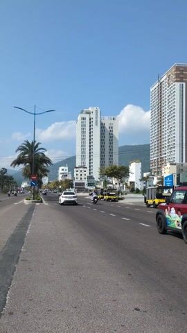 The video shows a street scene in a city, likely in Southeast Asia. A wide, multi-lane road stretches into the distance, with traffic including cars, motorcycles, and a small truck moving along it. On the left side of the road, there's a sidewalk with palm trees and streetlights. In the background, there are tall buildings, and mountains can be seen further in the distance under a blue sky with some clouds. The overall impression is of a busy urban environment on a sunny day.