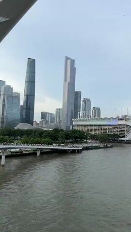 The video pans across a wide river with a cityscape in the background. On the river are several boats, some large and passenger-carrying, others smaller. Along the riverbank, there is a promenade with trees and people walking. In the distance, a large stadium with a white, curved roof is visible. Behind the stadium, a cluster of tall skyscrapers rises into the sky, including a distinctive, slender tower with a lattice-like structure, reminiscent of the Canton Tower. The sky is overcast and the overall lighting suggests either early morning or late afternoon. The water is a murky green-grey. The camera movement is smooth and steady, capturing the panoramic view of the city.