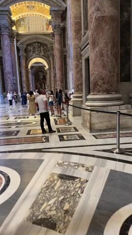 A wide shot from inside a large hall shows a group of people standing on a patterned marble floor. The hall has a high, ornate ceiling with gold and white decorations. Large columns line the sides of the hall, some with statues on them. The floor has black, white, and brown marble patterns. Many people are looking up at the ceiling and taking photos.