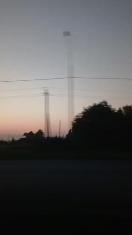 The video shows a view from a moving vehicle, likely a car, looking out a window. The scene depicts a rural landscape during either sunrise or sunset, with the sky displaying soft pastel colors transitioning from a light gray at the top to pink and orange near the horizon. Power lines are visible in the sky, running horizontally across the frame. The landscape below includes silhouetted trees and some structures that appear to be buildings or farmhouses, possibly partially obscured by mist or fog. The ground in the foreground is dark, suggesting it is a road or embankment. The motion blur indicates the camera is in transit. There are no people or distinct actions captured in the video, only the passing scenery.