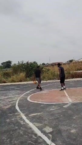 Two men are playing basketball on an outdoor court. The man on the left is wearing a black t-shirt and gray shorts, while the man on the right is wearing a black jersey and black shorts. The basketball court is located near overgrown vegetation. The sky is overcast. The man on the left is dribbling the basketball, and the man on the right is guarding him.