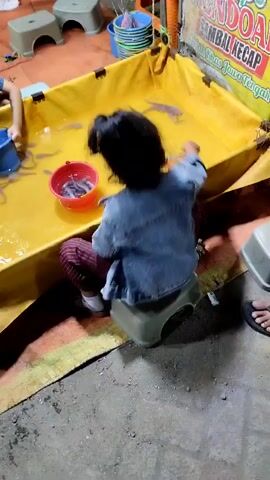 The video shows a close-up view of a shallow, yellow-lined pool filled with water and small fish. A young child, wearing a denim jacket and striped pants, sits on a small, beige stool next to the pool, holding a red bucket. The child appears to be engaged in fishing or playing with the fish. Other children and adults are partially visible in the background, suggesting a public gathering or event. 

The scene appears to be outdoors, possibly at a market or fair, given the presence of stalls and signs in the background. The lighting suggests it might be evening or a dimly lit area. One visible sign in the background contains text in Indonesian, which appears to be an advertisement for "Sambal Kecap" and "Bumbu Rasa Jawa Tengah" (Soy Sauce Chili and Central Java Flavor Seasoning).

The overall atmosphere seems relaxed and casual, with people enjoying a recreational activity. The focus is on the child's interaction with the fish in the pool.