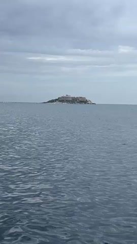 The video shows an island with a large, multi-story building on it, set in the middle of a large body of water, presumably the ocean. The sky is overcast with grey clouds, and the water is a dark blue with small ripples. The island appears to be rocky and somewhat barren, with the building dominating its landscape.  There is minimal human presence visible, if any. The video seems to be taken from a moving boat or vessel, as there are some subtle changes in the water patterns in the foreground.