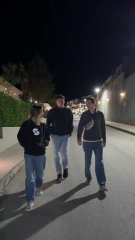 Three young adults are walking on a paved street at night. The woman on the left has long hair and wears a black hoodie with a logo on it and blue jeans. The man in the middle wears a black top and blue jeans. The man on the right wears a plaid shirt, a bag around his waist, and blue jeans. They are walking and talking, gesturing with their hands. The street is lined with buildings and plants. Streetlights illuminate the scene.