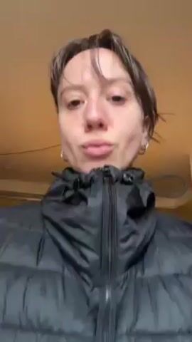 A person wearing a black puffer vest and a long-sleeved shirt is puckering their lips and turning their head back and forth, apparently looking at themself in a mirror. A Christmas tree is visible in the background, as well as a chair and other furniture. The background is somewhat cluttered. The person has short brown hair pulled back in a bun, and small hoop earrings. The lighting appears to be artificial and somewhat warm.