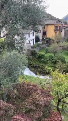 The video shows a scenic view of a small town nestled amidst mountains and a lake. The camera pans from a close-up of a small waterfall amidst lush greenery and houses. As the camera moves upward and outward, it reveals a panoramic view of the town with a variety of houses, gardens, and trees. In the background, towering mountains covered in green vegetation are visible, partially shrouded in clouds. The lake is a tranquil expanse of turquoise water. The town appears peaceful and quiet, offering a glimpse into a serene natural setting.