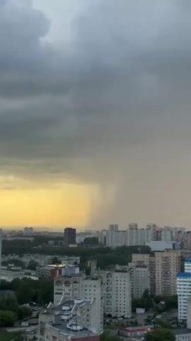 The video shows an aerial view of a city under a cloudy sky. A heavy rain shower is visible in the distance, appearing as a vertical curtain of water. The sun is setting, casting a warm yellow glow on the left side of the frame, contrasting with the dark, ominous clouds overhead. Several buildings of varying heights are scattered throughout the cityscape, with a few taller structures in the mid-ground and foreground. The overall atmosphere is dramatic, with the impending rain and the fading sunlight creating a sense of impending change.