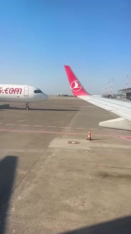 The video shows an airport tarmac on a clear, sunny day. Two airplanes are visible. The airplane on the left is white with red lettering that reads "flypgs.com". The airplane on the right has a pink tail with a red logo. The wing of this airplane is in the foreground of the shot. A traffic cone is visible on the tarmac between the two planes. Other airport structures, including what appear to be jet bridges, are visible in the distance. The sky is a clear blue.