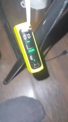 A yellow electronic device with a digital display is shown. A white cable is plugged into the top of the device. The display shows the number "00", a green bar graph with three colored segments (red, blue, and green), and the number "3". A small green light is also illuminated below the number "3". The device appears to be attached to a black metal object, possibly a chair or table leg, and rests on a dark wooden floor.