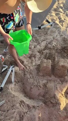 A person wearing a straw hat and patterned shorts is pouring water from a green bucket into a hole in the sand, creating a muddy puddle. The person is barefoot and standing on a sandy beach. There are sandcastles in the background. The scene is illuminated by natural light.