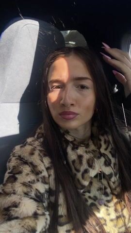 A woman in a leopard print coat and long dark hair is sitting in the passenger seat of a car. A man with curly dark hair sits next to her. Music is playing. The woman smiles and closes her eyes. The car appears to be stopped.