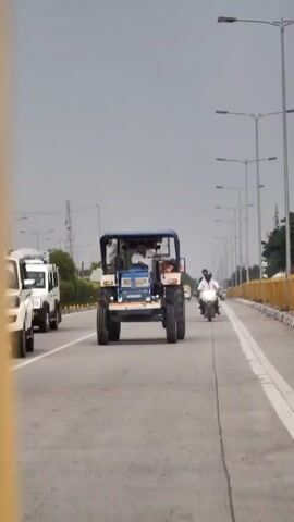 A white SUV is driving on a highway next to a blue tractor. There are streetlights lining the right side of the highway and a yellow barrier beyond them. A person is driving the tractor and another person is riding on the back. Two motorcycles are also visible on the highway, one of which is passing the tractor. The sky is cloudy.