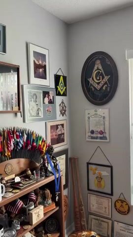 This video shows a corner of a room with walls covered in framed pictures, certificates, and other memorabilia. The items include photographs, what appear to be awards or degrees, and a large circular emblem featuring a compass and square, suggesting a connection to Freemasonry. A bookshelf is visible on the left, filled with more items and decorated with numerous small flags. The overall impression is of a highly decorated space, possibly a personal study or office, filled with collected items and achievements.