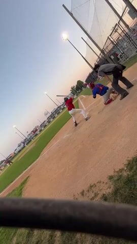 A baseball game is in progress as the sun begins to set. A batter wearing a red jersey and white pants swings at a pitch. A catcher in blue and red gear is behind home plate, and an umpire in a grey shirt and black pants stands to the side. The baseball field is visible, with a dirt infield and green outfield. Lights illuminate the field. In the background, a fence and spectators can be seen. The sky is a gradient of blue and orange, indicating dusk. The camera is positioned low, giving a perspective from the dugout or bleachers.