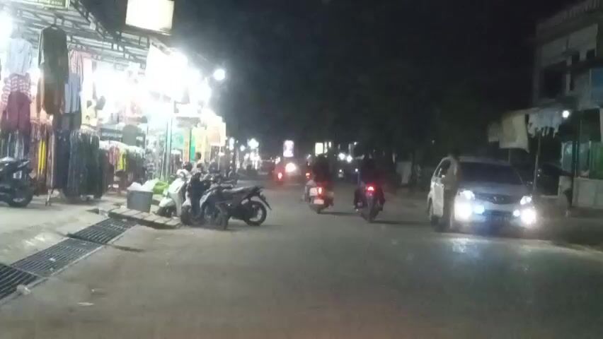 The video shows a street scene at night, possibly in Southeast Asia. On the left, there are several stalls displaying clothing, illuminated by bright lights. Several motorcycles are parked in front of the stalls. On the right side of the street, there is a white car with its headlights on, and a person standing next to it. Further down the street, several other motorcycles are in motion, their taillights visible. The street is paved and appears to be in a commercial or residential area. The overall atmosphere is active with ongoing traffic and commercial activity despite the darkness.