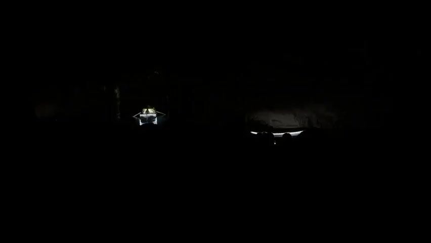 The video shows two boats navigating a dark, cave-like environment. The boat on the left has a yellowish light illuminating its interior, while the boat on the right is darker, with only a few faint white lights visible. The boats are the only objects visible. The walls of the cave are dark and undefined, except for the areas where the boat lights reflect against them. The boat on the left seems to be approaching the viewer, while the boat on the right may be moving away. The overall atmosphere is mysterious and dimly lit.