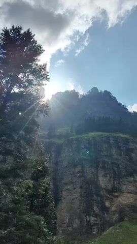 The video pans across a mountainous landscape. In the foreground, a tall evergreen tree is on the left, with its branches obscuring part of the view. The sun is visible through the clouds, creating lens flare and rays of light that illuminate the scene. The mountains in the background are steep and rocky, with patches of green grass and trees growing on their slopes. The sky is blue with scattered white clouds. The overall impression is one of a serene and majestic natural environment.