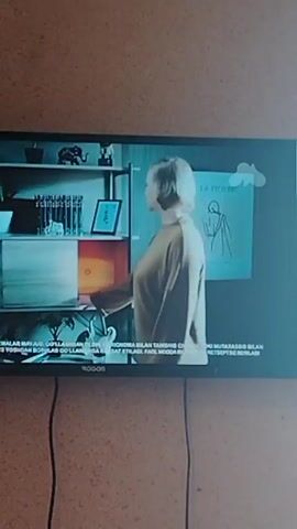 The video shows a TV screen displaying a medical animation. Initially, a red MIG logo is visible on a blurred background. The screen then shows a glowing blue outline of a human figure, with internal organs in red. A blue circular clock graphic appears around the figure, with clock hands moving around the perimeter. Text in a foreign language is visible at the bottom of the screen. The TV itself is black and is mounted on a wall with a textured tan or light brown surface. Two black power cables hang down from the TV.
