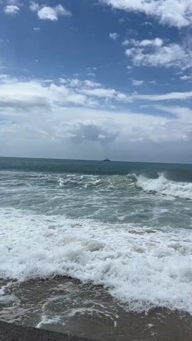 The video shows the ocean on a partially cloudy day. Small waves crash onto the shore, creating white foam. In the distance, a boat or structure is visible on the horizon. The sky is blue with scattered white clouds, and some darker clouds are also present.