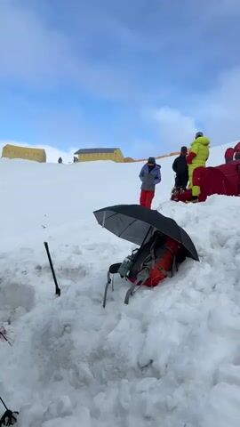 The video shows a snowy mountainside with several red tents set up. In the foreground, a backpack is partially covered by an open black umbrella. Several people in winter clothing are visible on the slope, some near the tents and others walking uphill in a line. In the background, there are yellow structures and more tents. The sky is cloudy with patches of blue.